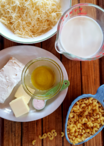 all the makings for homemade Mac and cheese - butter, cream cheese, cheese, milk and pasta on a wood cutting board